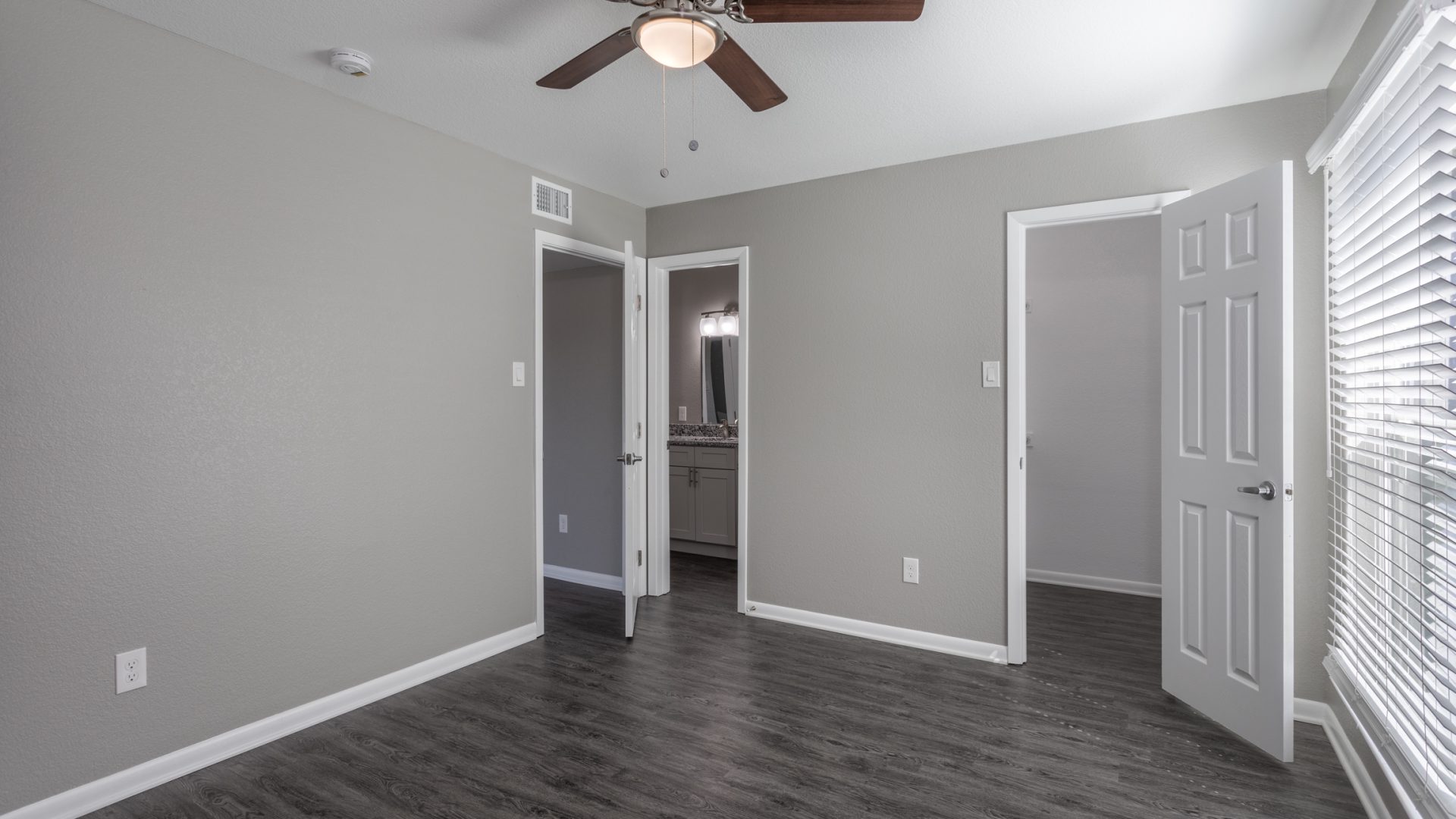 Picture of a bedroom in an apartment with dark brown, vinyl flooring, gray walls, and a ceiling fan.